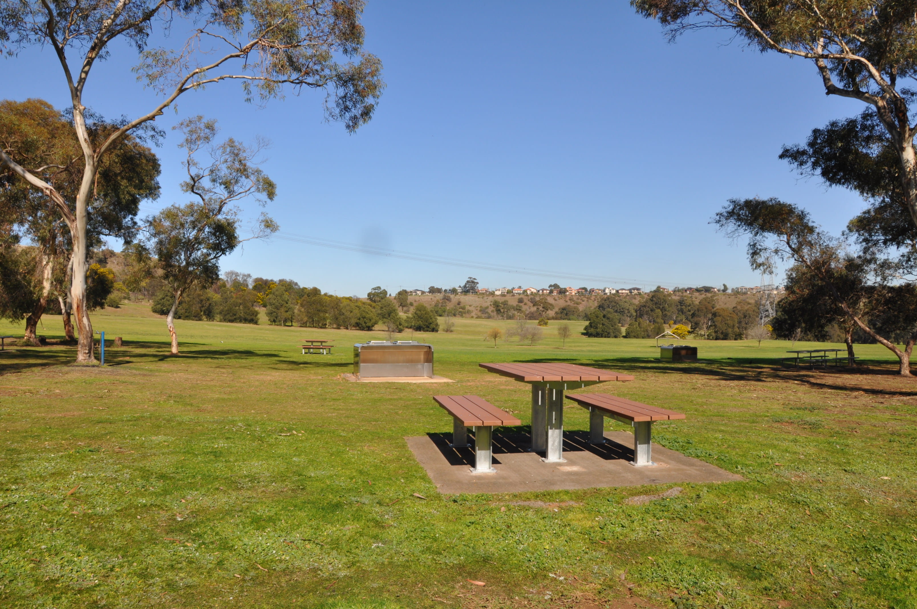 New picnic tables and barbecues at Brimbank Park. They are spread out over a grassy area, with trees and a blue sky in the background.