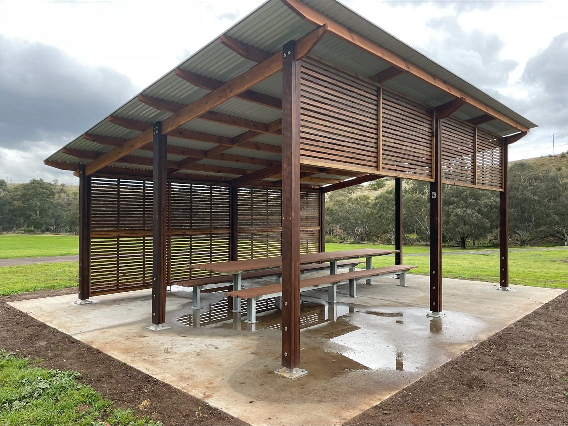 A new shelter, made from timber and steel, has been built at Brimbank Park. Underneath it is a large concrete slab and two long picnic tables. It is surrounded by grass and trees.