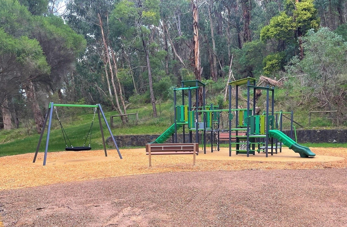 A new playground, with a swing basket on the left, and typical climbing equipment and slides on the right. The equipment is blue and green in colour, and sits on top of tanbark. Bushland is in the background.