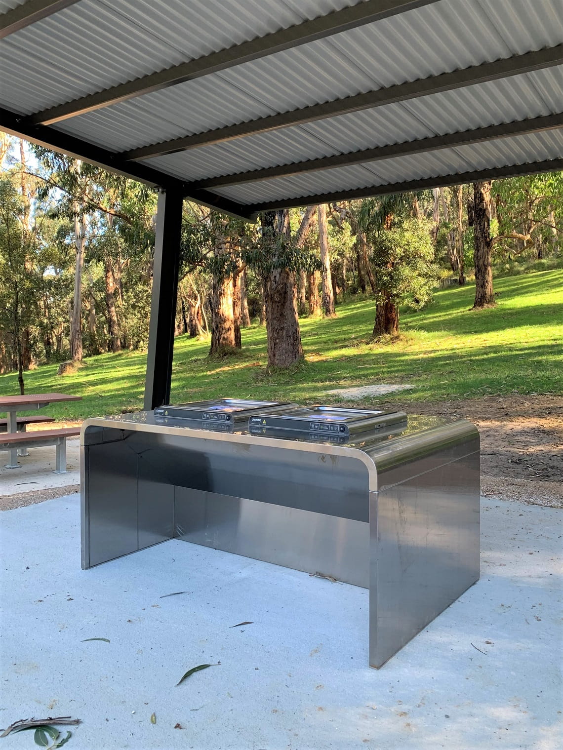 A stainless steel electric barbecue sits on top of a newly-poured concrete slab. The barbecue is under an open shelter with a corrugated iron roof.