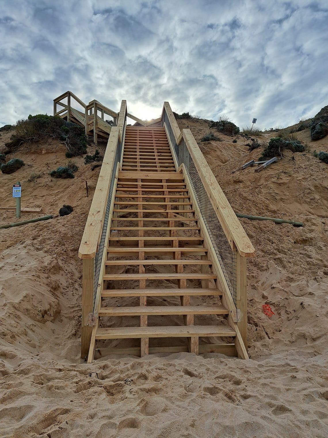 A portrait photo of the new stairs at Number 16 Beach. The photo is taken from the bottom of the stairs, looking up from the beach to the top of the stairs, which leads to grassy sand dunes. 