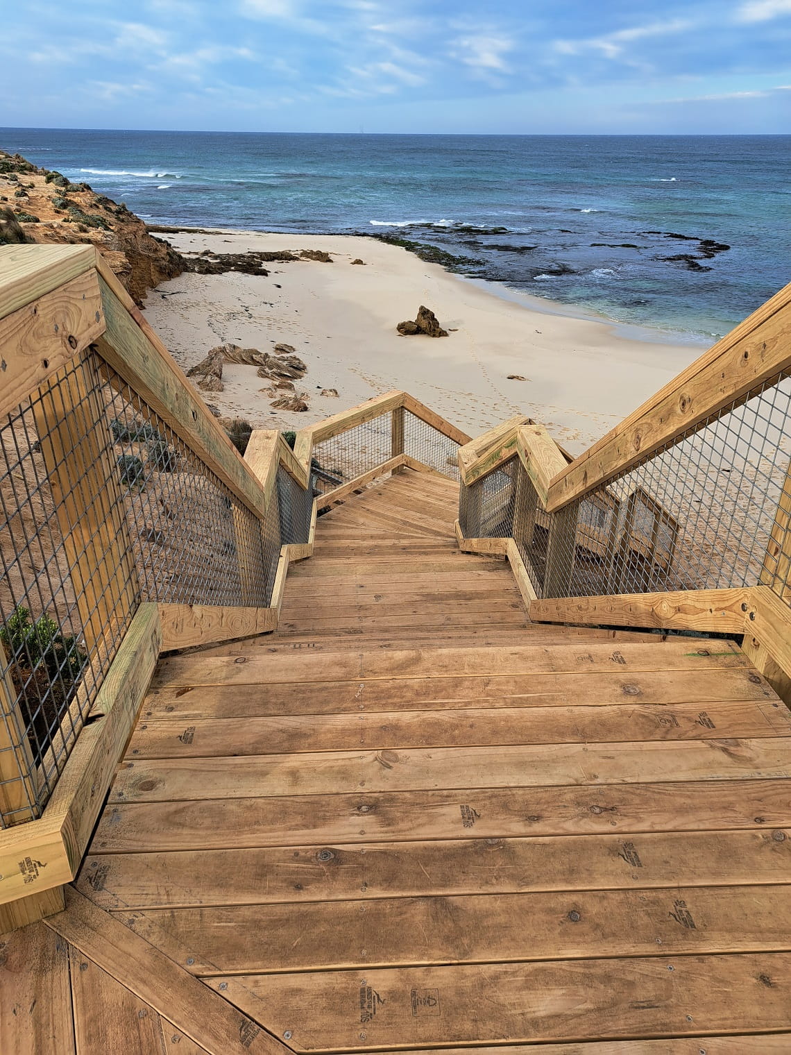 A portrait photo of the new stairs at Number 16 Beach. The photo is taken from the top of the stairs, looking up down the stairs onto the beach. The water and blue sky is in the background. 