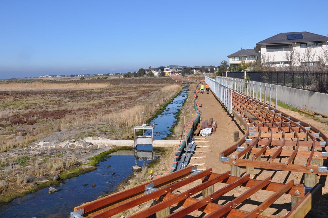 The construction of the Bay Trail extension. A wooden frame is shown travelling from the front of the image into the distance, alongside wetlands and double-storey houses.