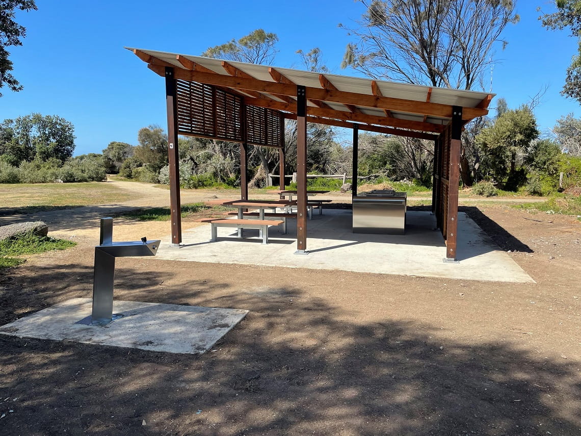 A new shelter sits on top of a new concrete slab, in the sun. A drinking fountain is in the foreground. 