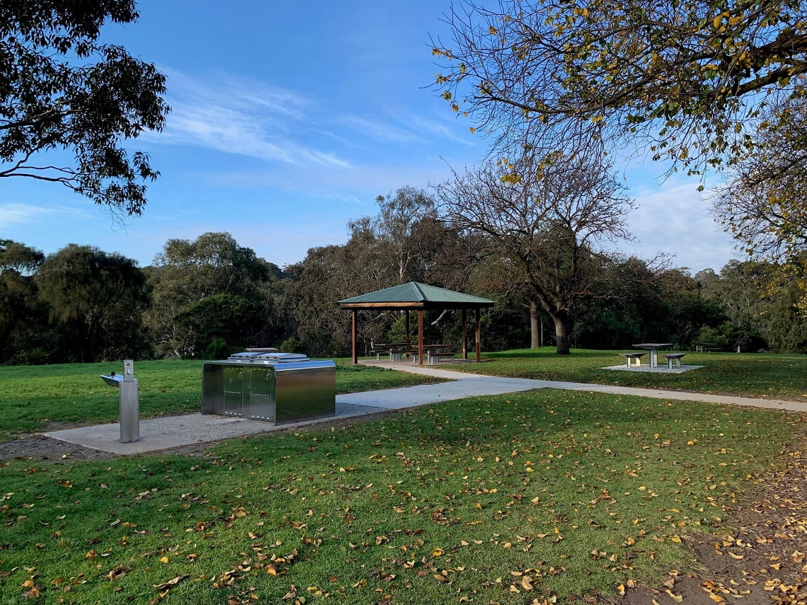 New barbecues, drinking fountains and picnic tables are arranged on a flat grassed area among deciduous trees with shedded and autumn leaves. 