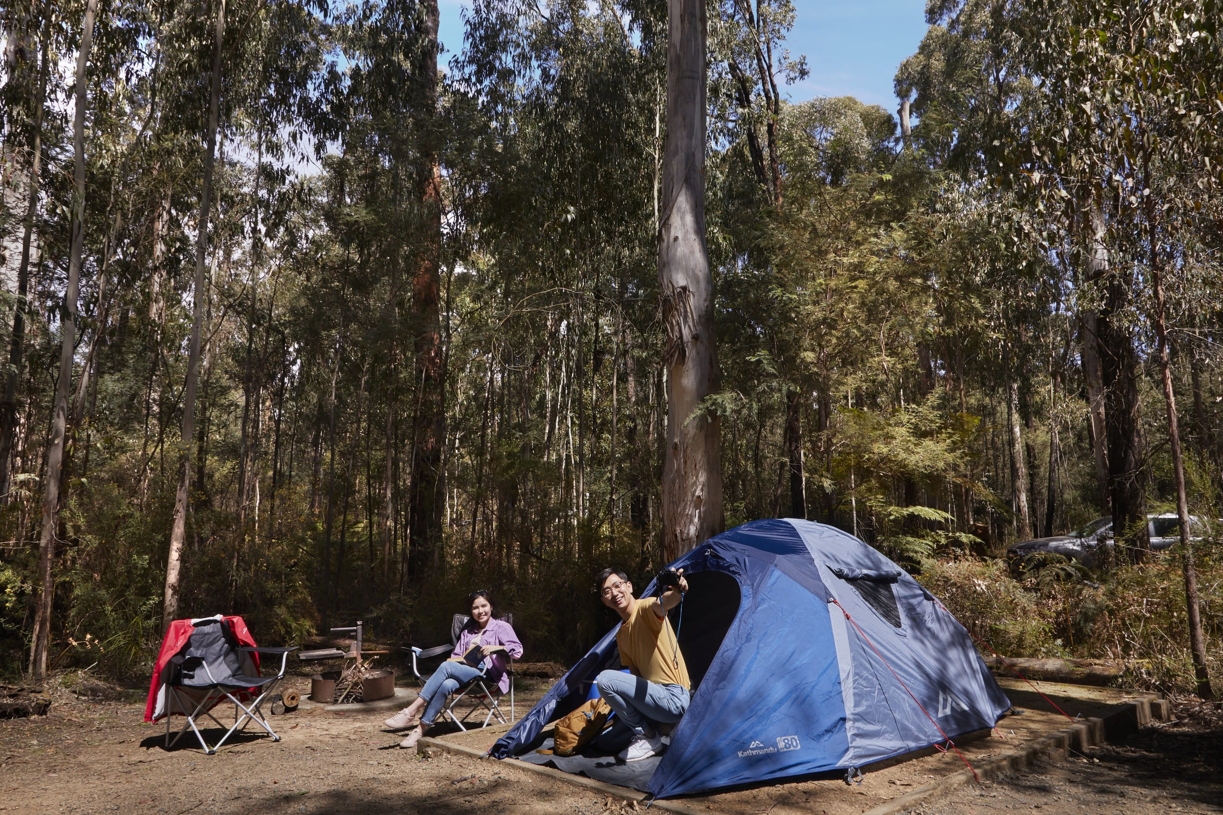 Two people near a blue tent with tall forest in background