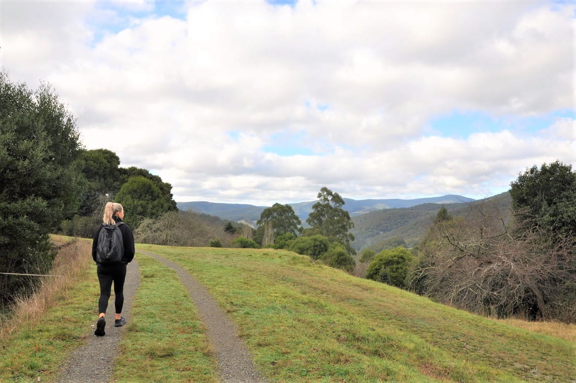 A blonde girl in black clothes walking along a grassy track, with mountain ranges in the background.