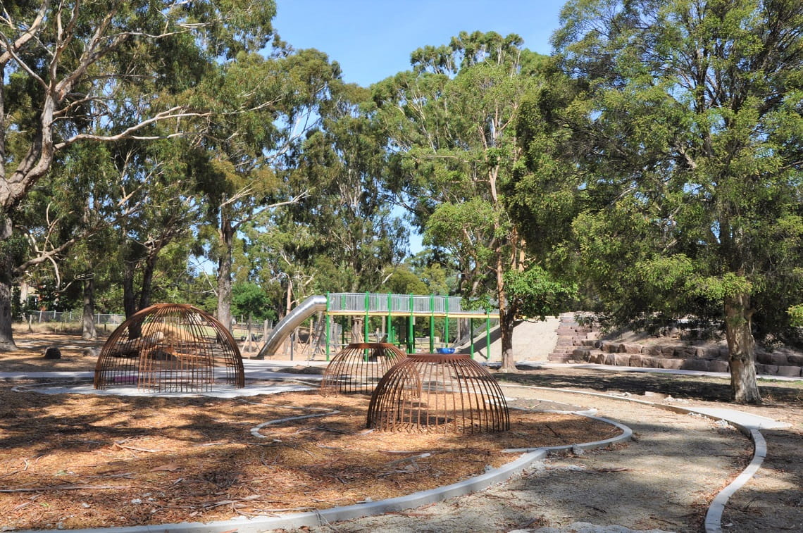 A distant view of the Wattle Park playscape currently under construction. Climbing equipment can be seen in the foreground, with an unfinished path winding towards the green and yellow frame of the tram fort in the distance. 