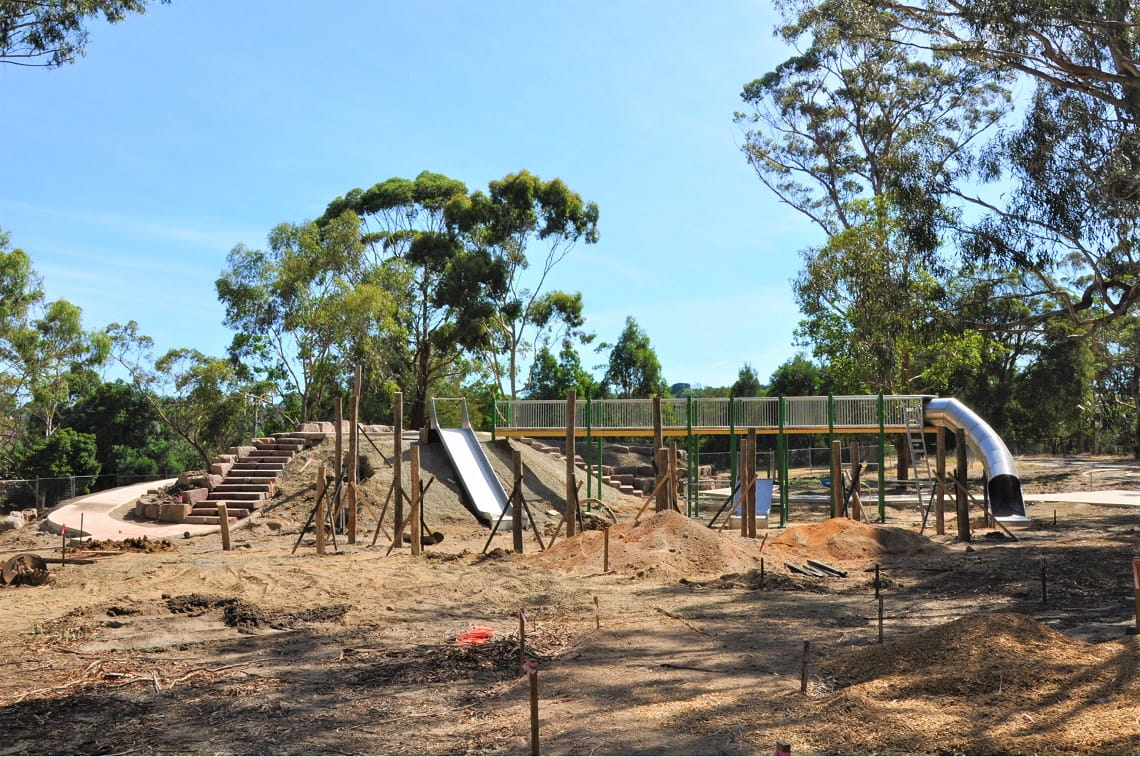 The Wattle Park playscape is under construction. To the left of the image is a mound of carefully placed rock, with a pathway winding up to level two of the playscape. The playscape includes slides and a tram fort. Trees and disturbed earth from construction surround the playscape. 
