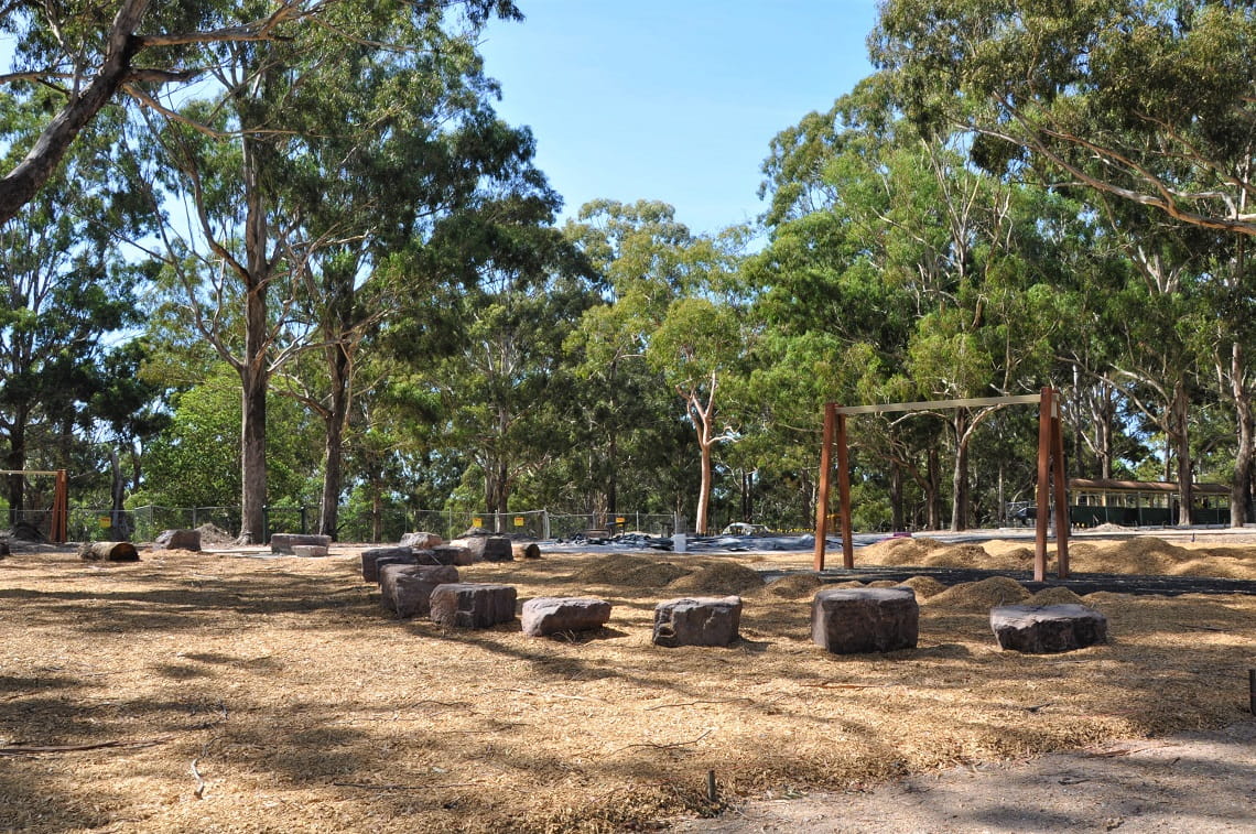 The structure of a swingset is in place, surrounded by large rocks for seating. The new playground equipment sits underneath the dabbled shade of tall gum trees. 