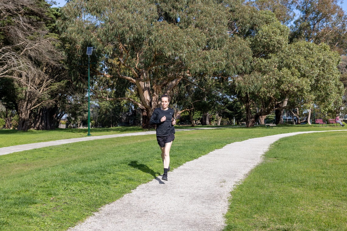 A young woman with dark hair in black clothes is running on a light grey track through the centre of the image. Either side of her is green grass, and in the background are tall trees. 
