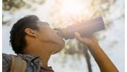 A low angle photo of a man drinking water from a bottle wearing casual clothes and standing against trees on a sunny day