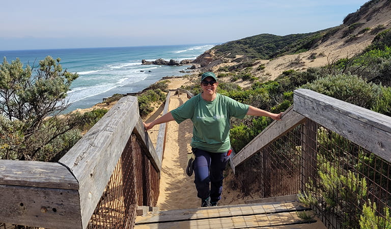 A woman wearing a Parks Victoria volunteer top walking up stairs with a beach in the background.