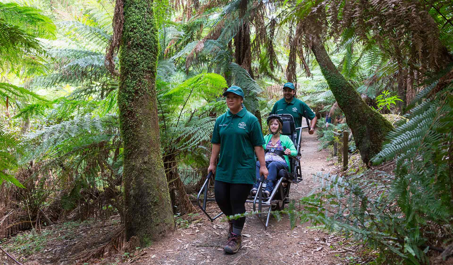 Two people assist a third person in an all terrain wheelchair along a track surrounded by nature