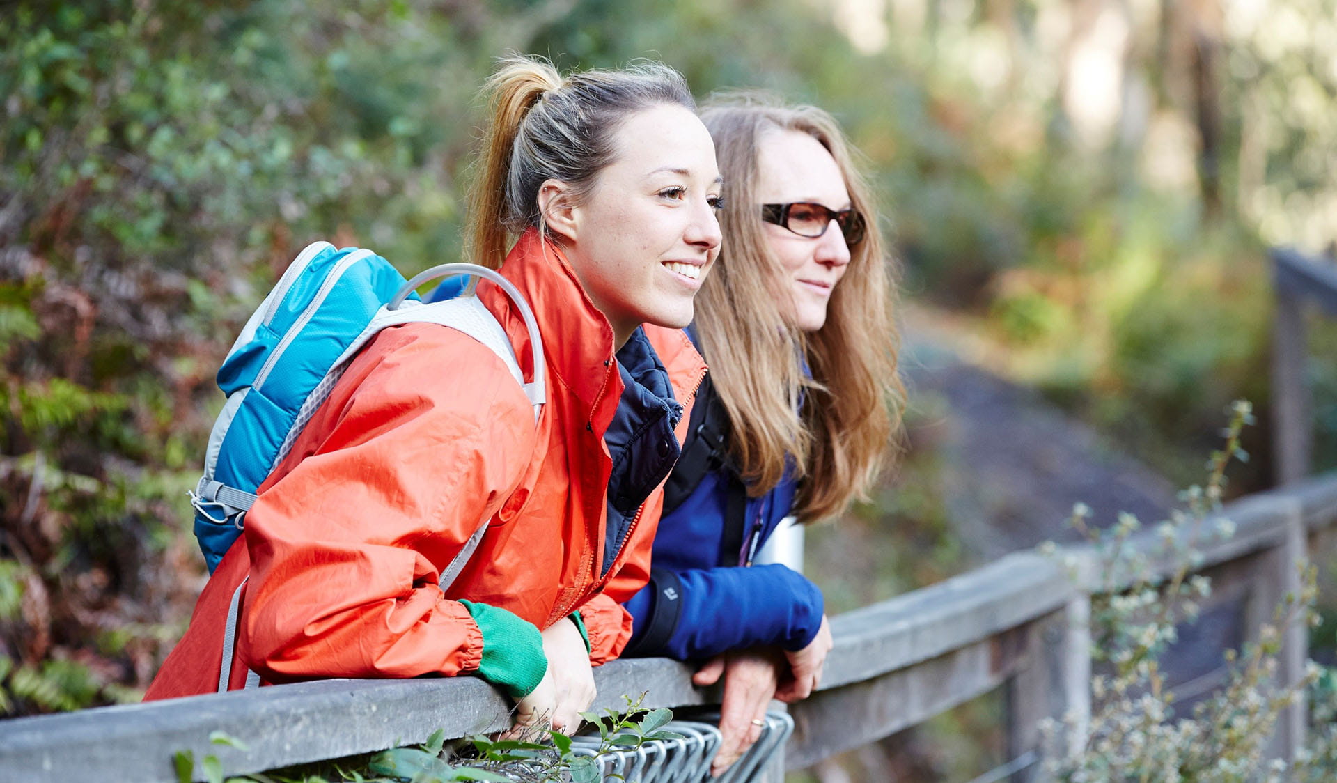 Two women in colourful rain jackets smiling as they lean against and look out over a wooden safety barrier