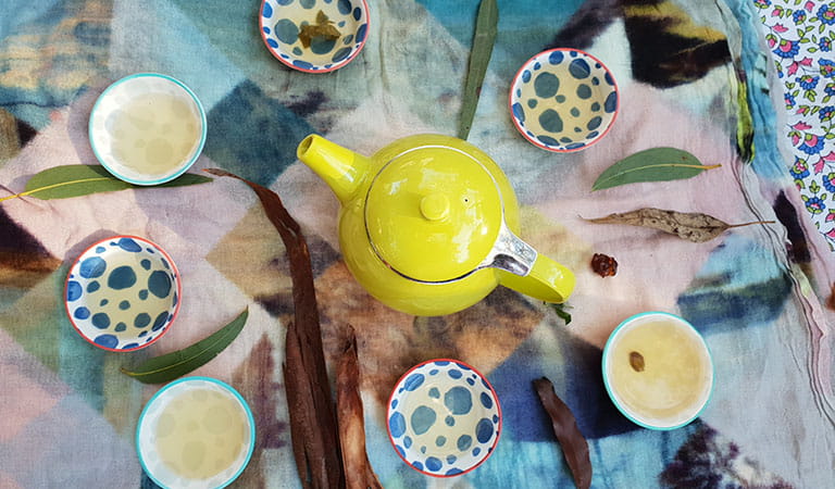 Overhead view of a yellow teapot and cups of tea on a colourful tablecloth.