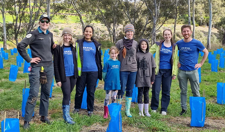A group of smiling adults and kids with a Parks Victoria ranger posing between planted trees.