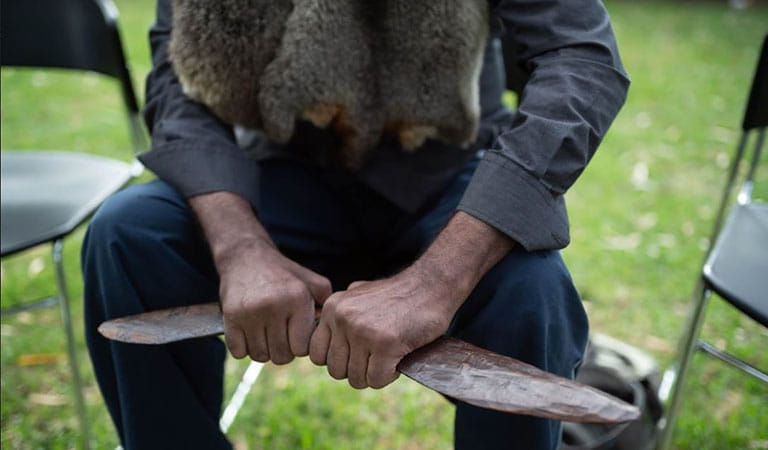 Close up of a seated person wearing native animal fur and holding a carved wooden tool in their hands.