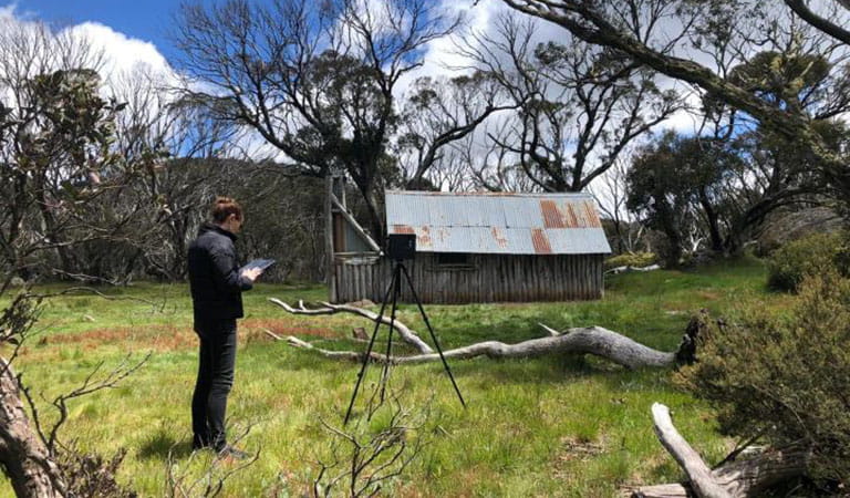 A person standing in front of an alpine hut with 3D modelling camera equipment.