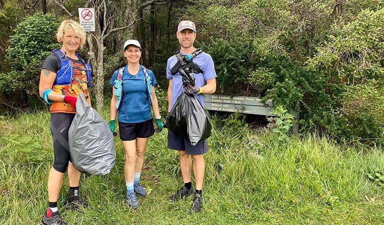 Two women and a man in exercise clothing and gloves standing on grass, holding bags of collected rubbish.