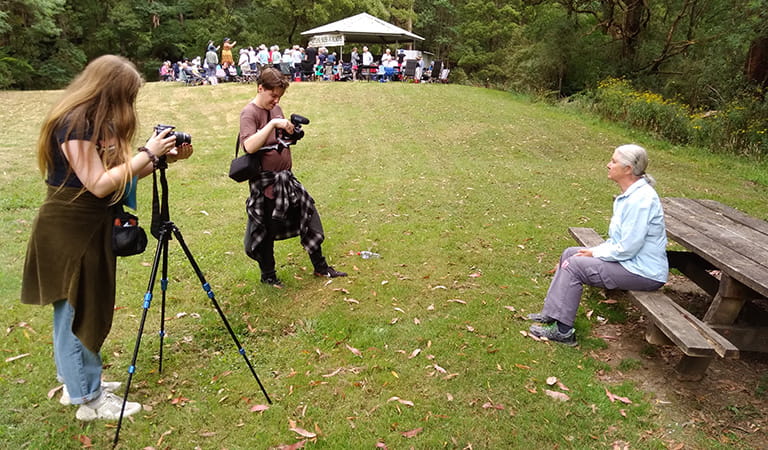 Two people with cameras filming an older woman sitting on a park bench, with a group of people gathered in the background.