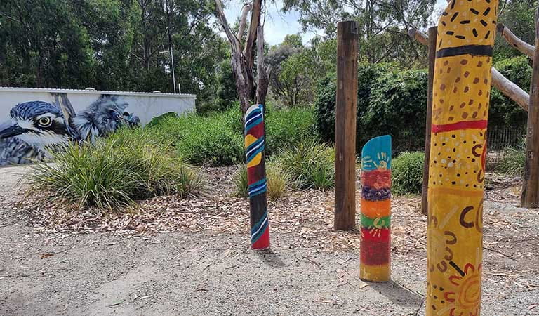 Colourfully decorated upright logs of wood, with a bird mural on the wall of a structure in the background.