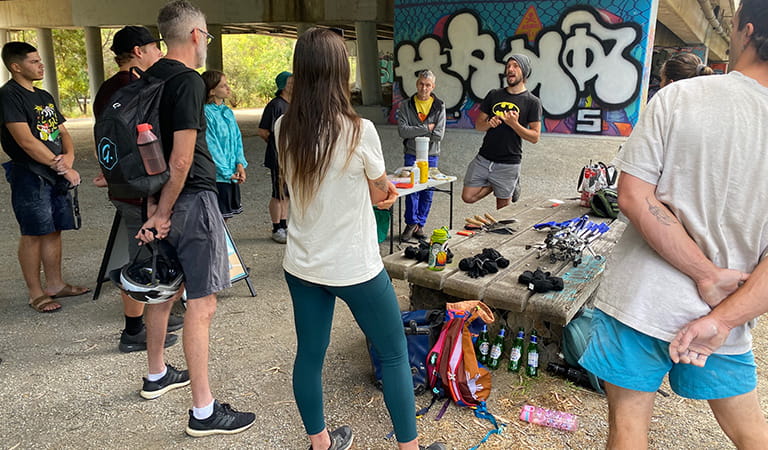 A group of people standing under a bridge, listening to a presenter, with a graffiti wall in the background.