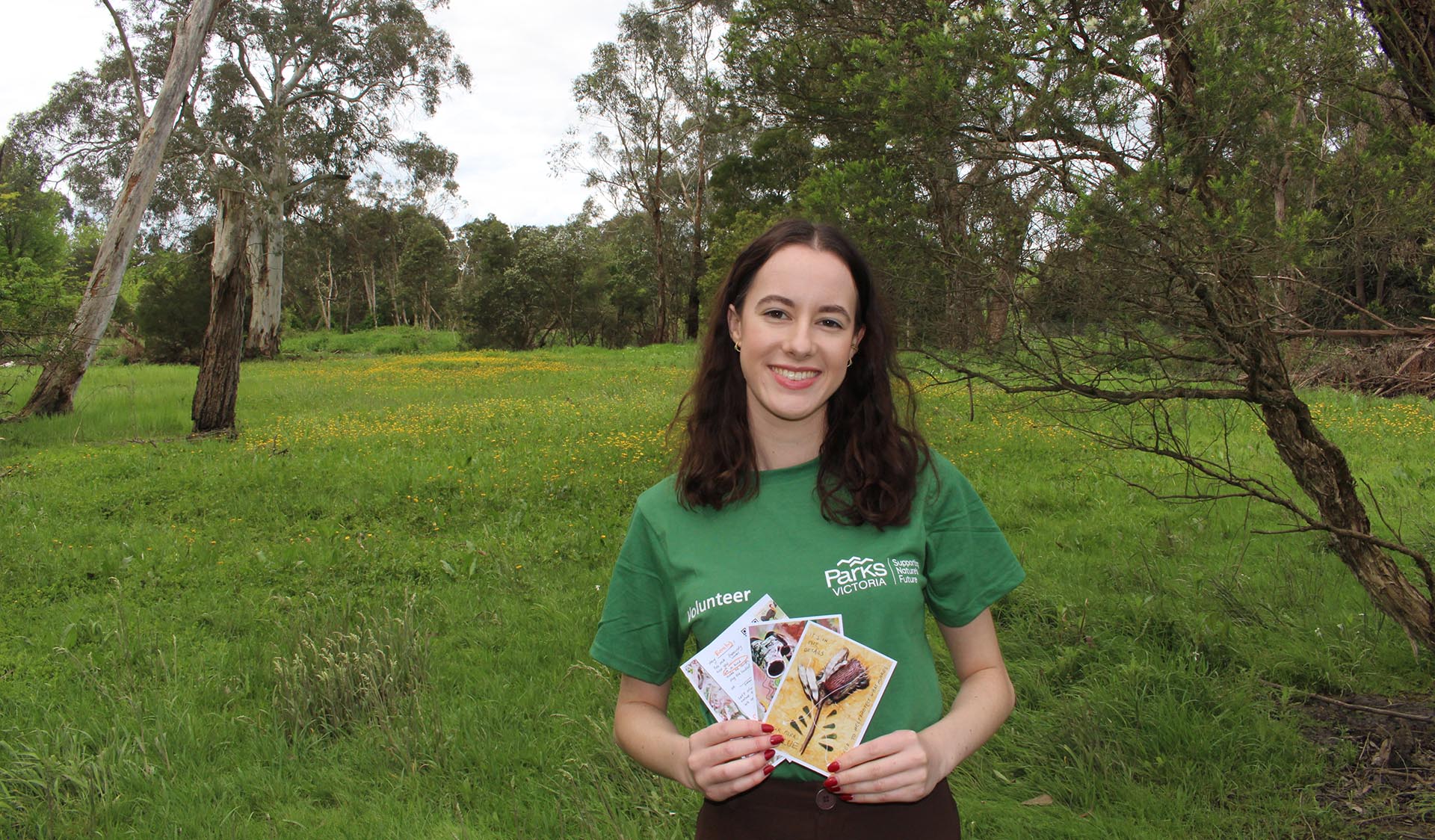 A girl wearing a Parks Victoria volunteer t-shirt, smiling and holding up a set of illustrated cards with grass and trees in the background.