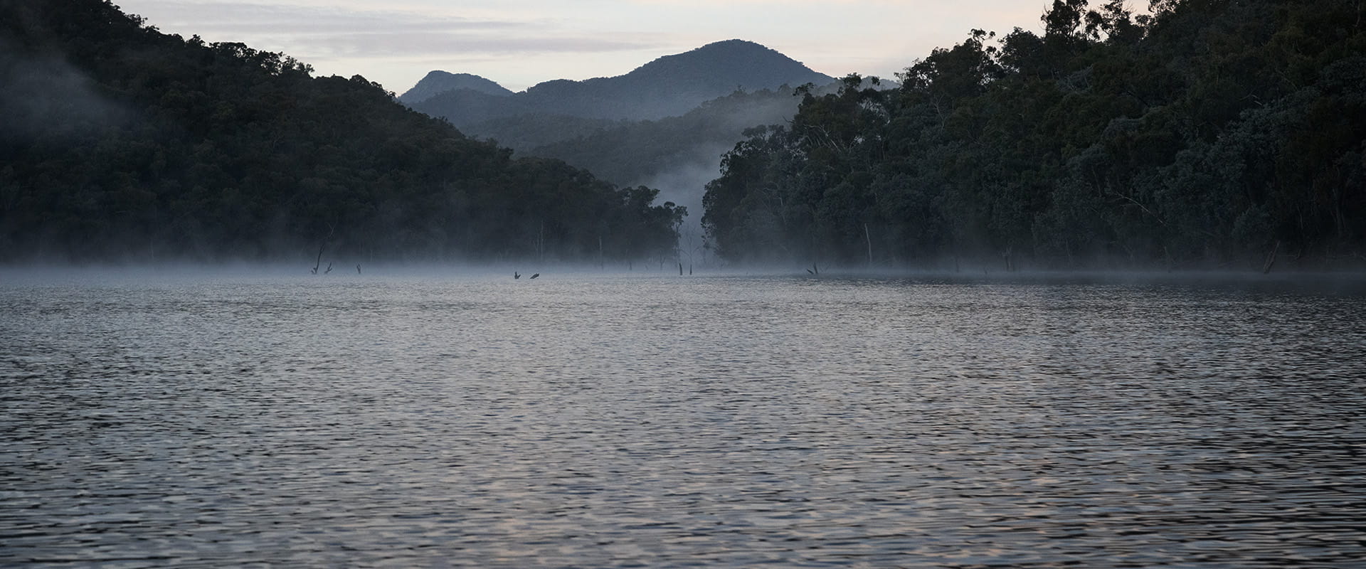 Mist rises on a lake in the foreground. Forested mountains rise in the background and far off in the distance. 
