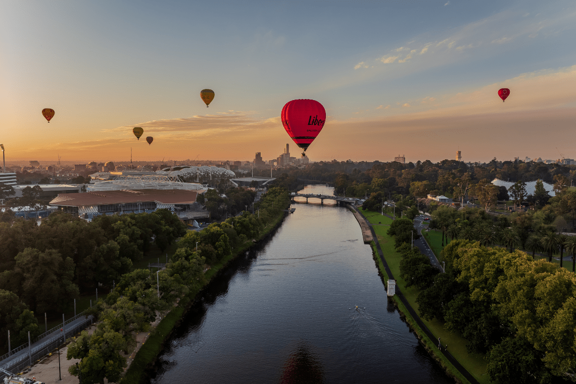 Hot air balloons floating over Melbourne at sunrise, with the Yarra River, sporting precinct, and city skyline visible beneath a golden sky.