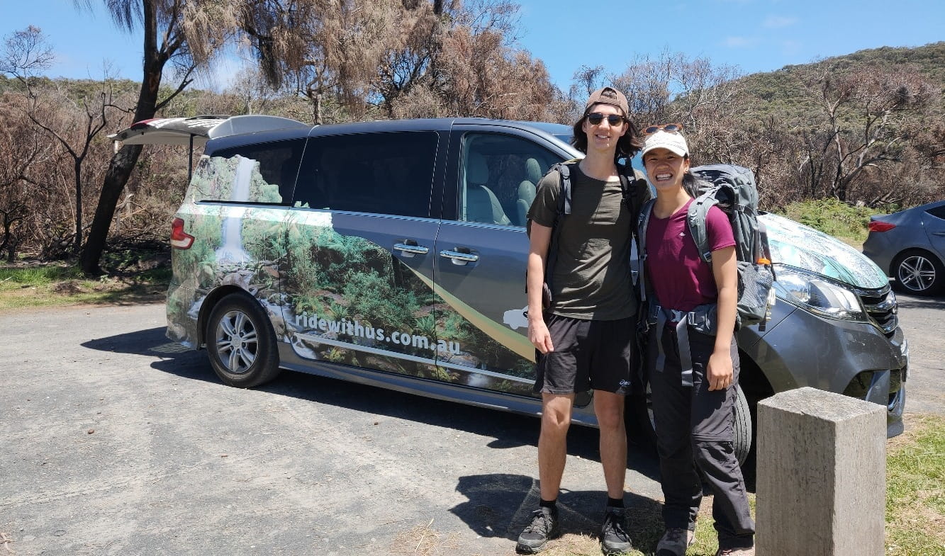 A smiling couple stands in front of a Ride With Us transport van. 