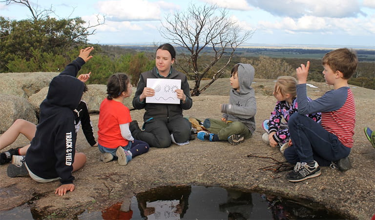 A woman in a ranger uniform presenting illustrations to a group of children as they sit on a large boulder.