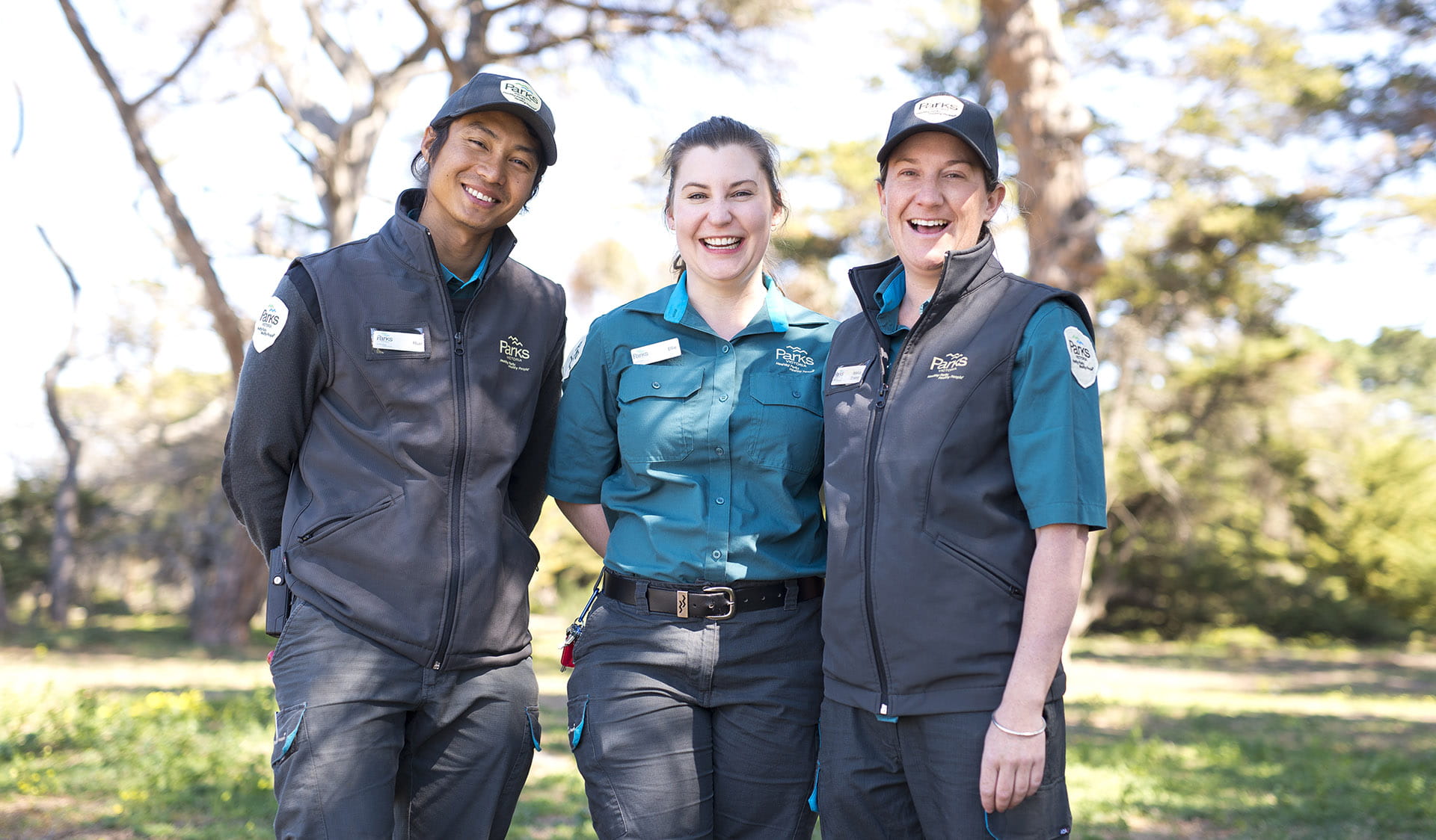 Three rangers in Parks Victoria uniform smiling at the camera with trees in the background.