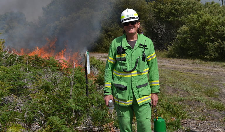 A man in a bright green uniform poses while supervising a planned burn in the background.