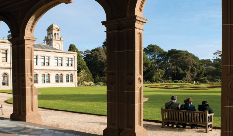 Looking out through sandstone pillars onto the lawn and mansion facade, with three people sitting in a park chair at the edge of the lawn.