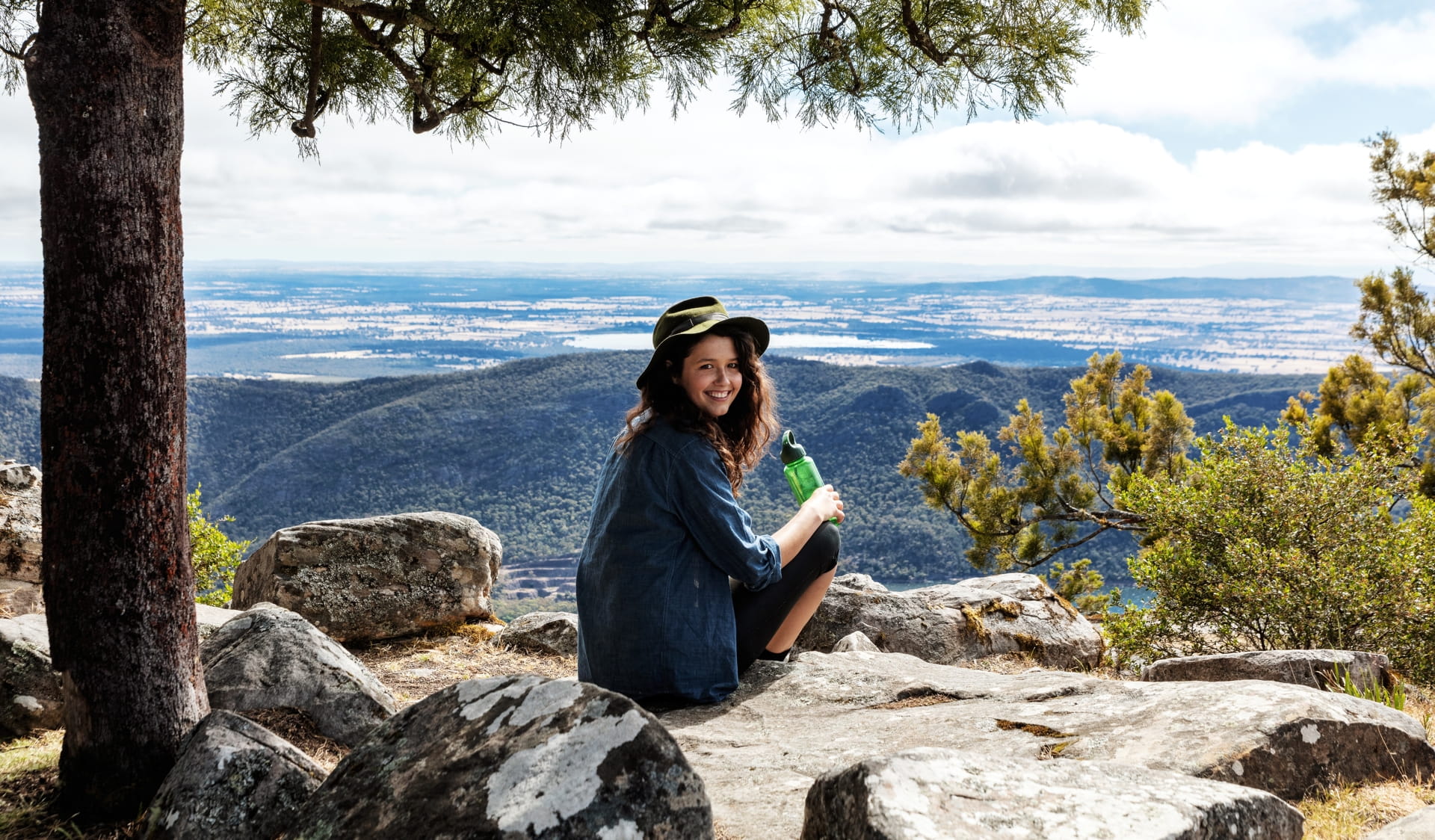 A young volunteer with long dark hair and a sunhat sits overlooking a mountain vista. 