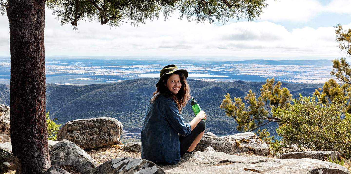 A smiling female park visitor wearing a hat and holding a water bottle, sitting in nature overlooking a view.
