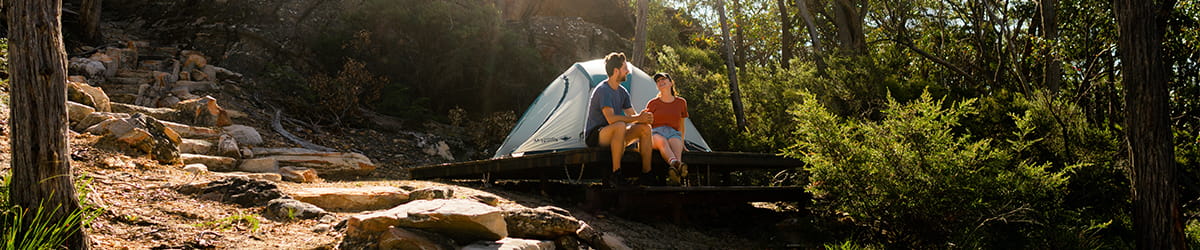 A man and woman sitting on a tent pad outside their tent, surrounded by nature.