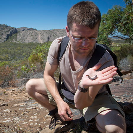 Man crouched looking at a small lizard perched on his open palm.