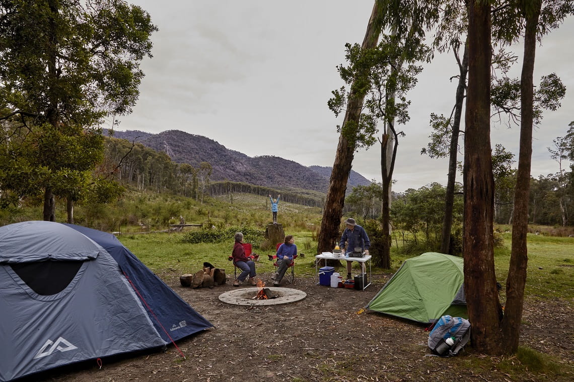 Picture shows a large family group of five people camping outdoors with two tents, a large one and a small one, with the adults of the group gathered around a fireplace with fire.