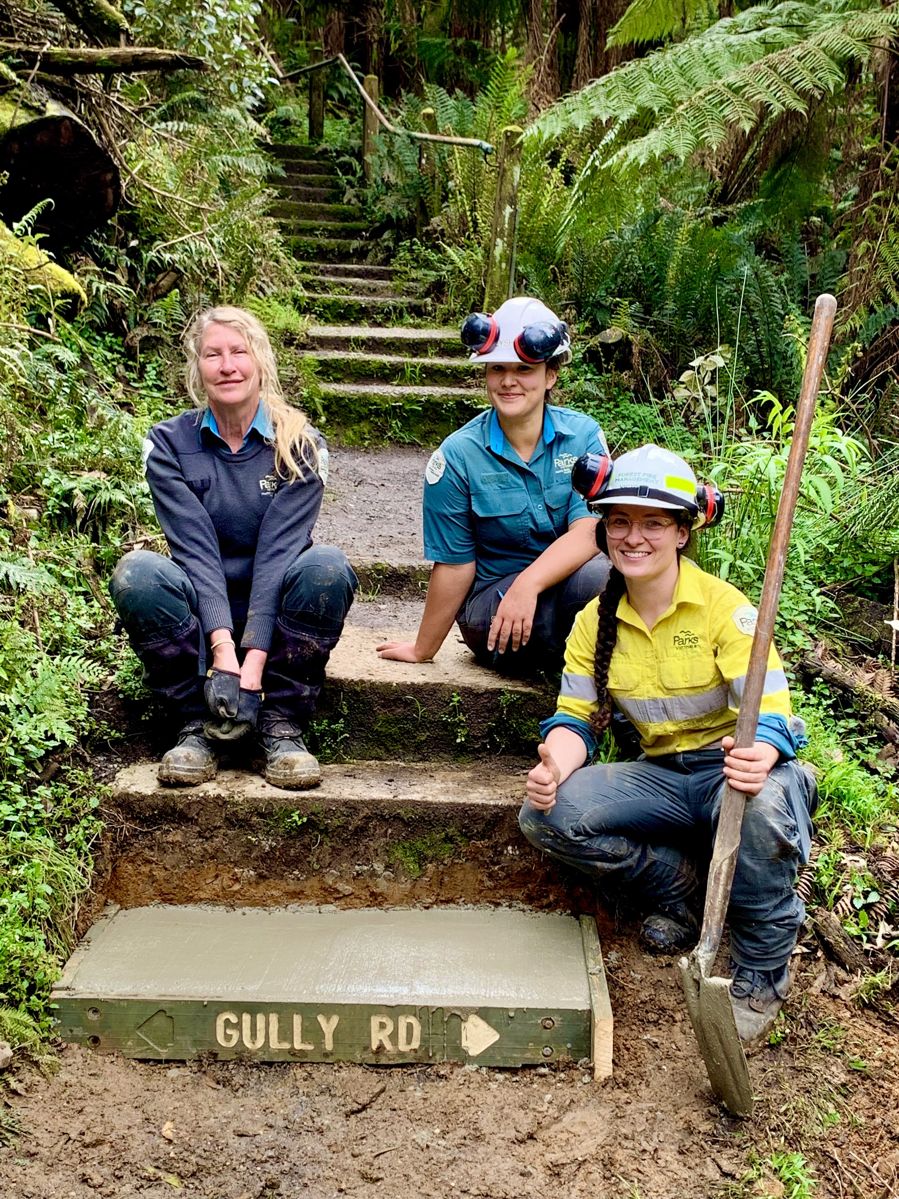 Shows a new concrete step in place on a freshly cleaned walking track  surrounded by forest. Three women sit on the steps, one is a park ranger with working gloves and two others are in high viz with overalls and hard hats, all are smiling at the camera with a sense of a job well done.