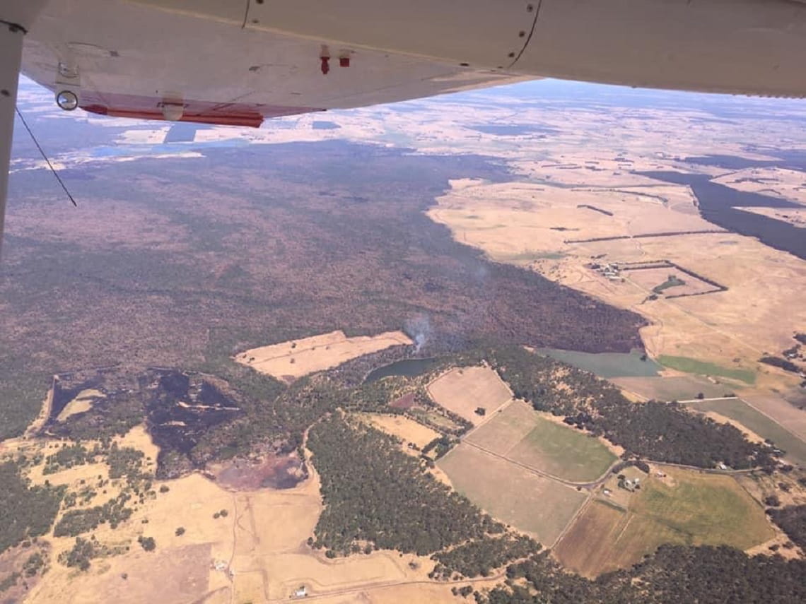 View from from the window of a fixed wing plane looking over a forested area adjoining paddocks. There is a smoke column from a fire in the distance