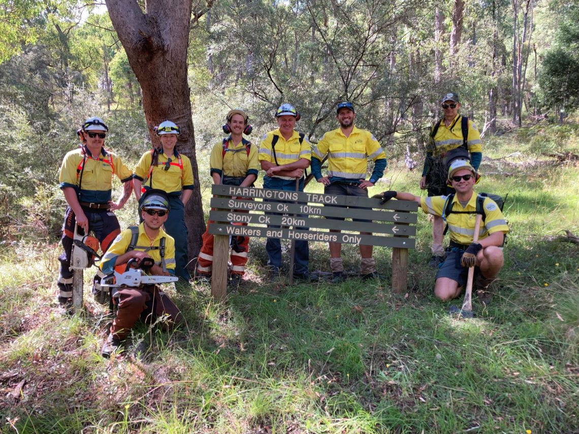 A group of eight people in high-vis uniforms, carrying cutting tools, in front of a Harrington's Bridle Track sign.