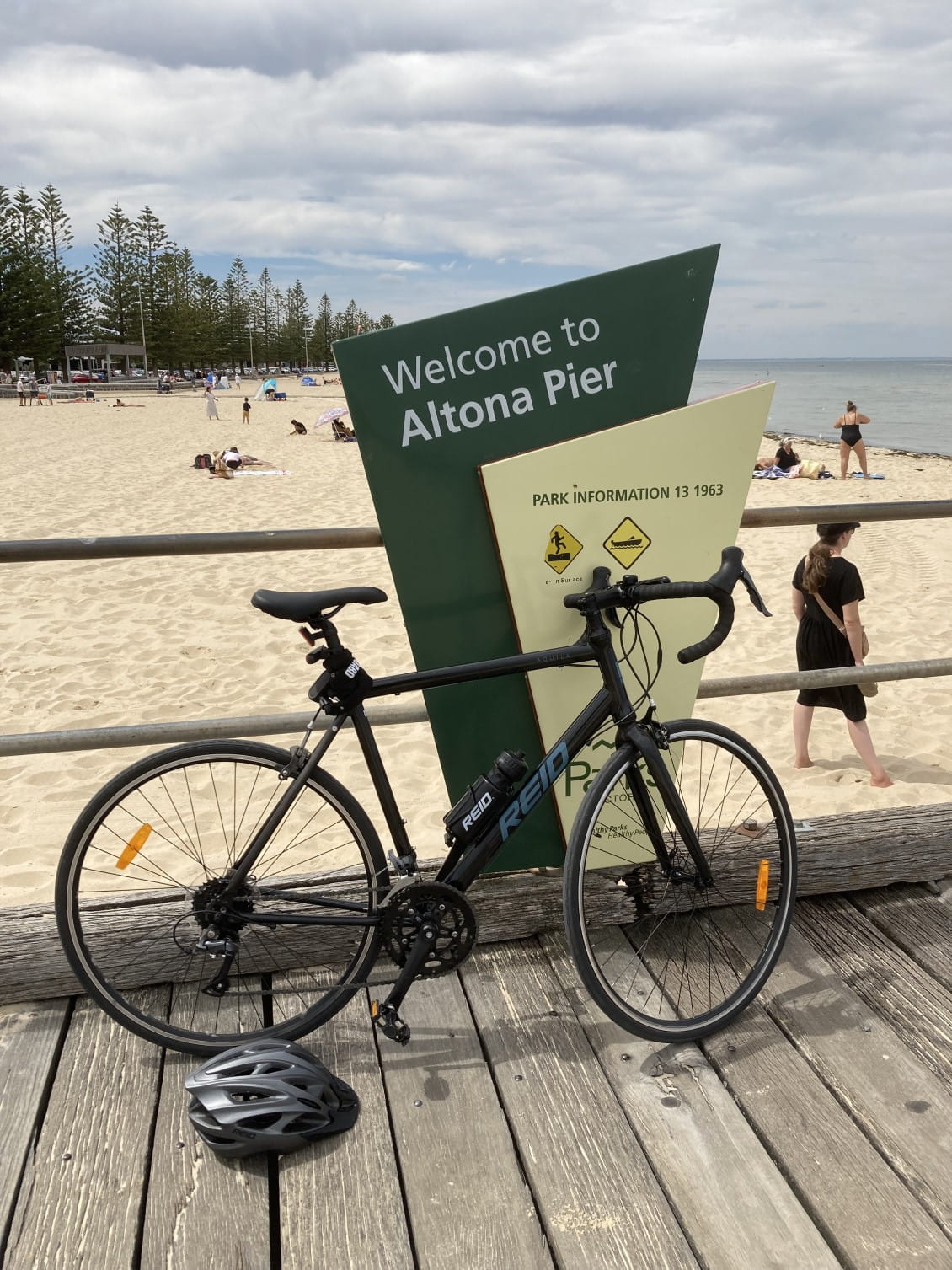 A bike leans against a railing with a sign saying "Welcome to Altona Pier". Behind the railing is a sandy beach with people sunbathing.