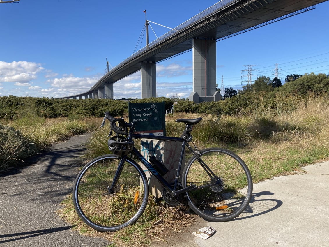 A bike leans against a sign saying "Welcome to Stony Creek Backwash". Behind them, the towers of the Westgate Bridge.