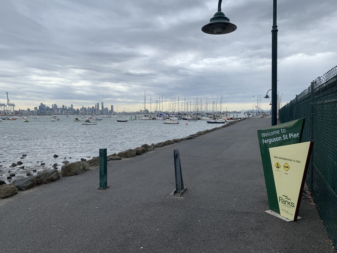 A sign saying "Welcome to Ferguson Street Pier". Behind it a pier stretches out into the water with boats moored alongside.