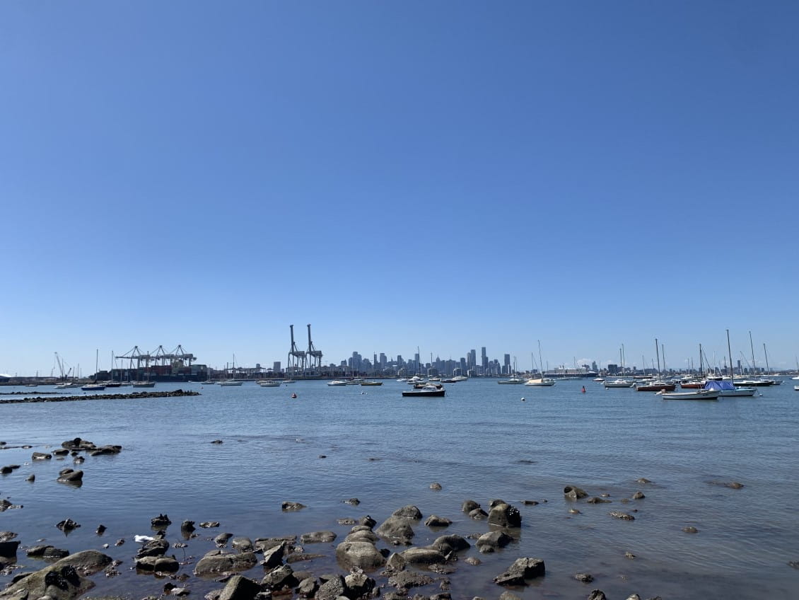 An expanse of water with boats. Beyond them in the distance are the cranes of a dockyard and beyond that the towers of central Melbourne.