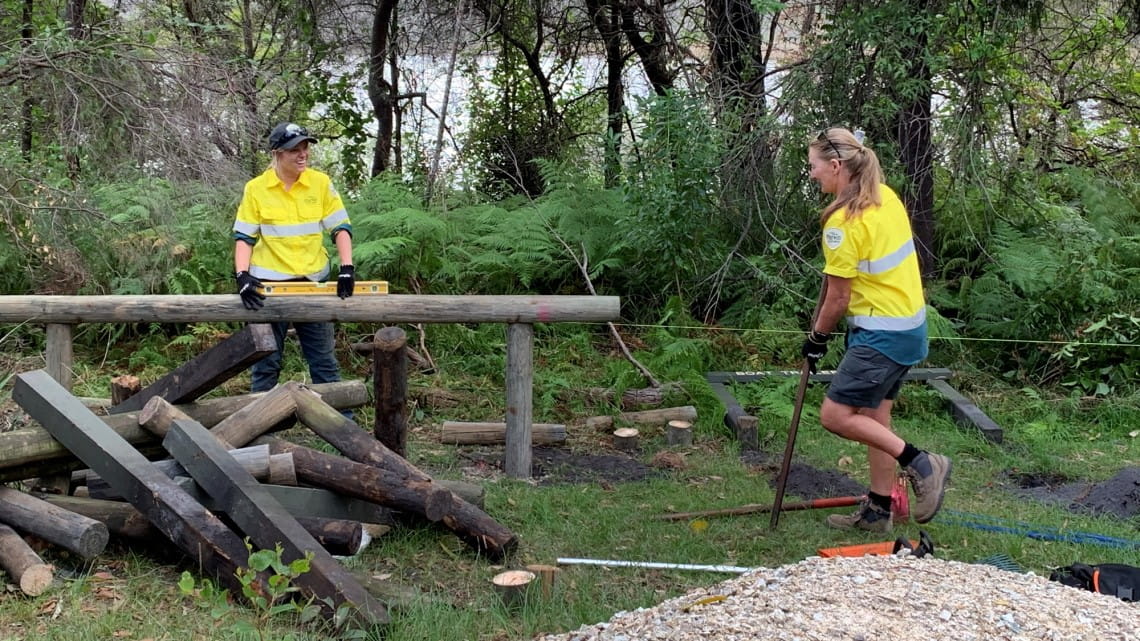 Two women work with tools on a wooden fence surrounded by forest