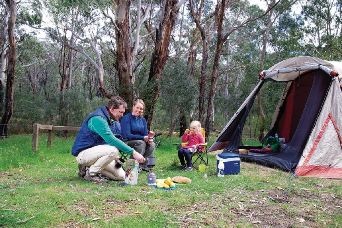 Picture shows a family of Mum, Dad and a young child out the front of their tent, warming themselves around a small camp fire. 