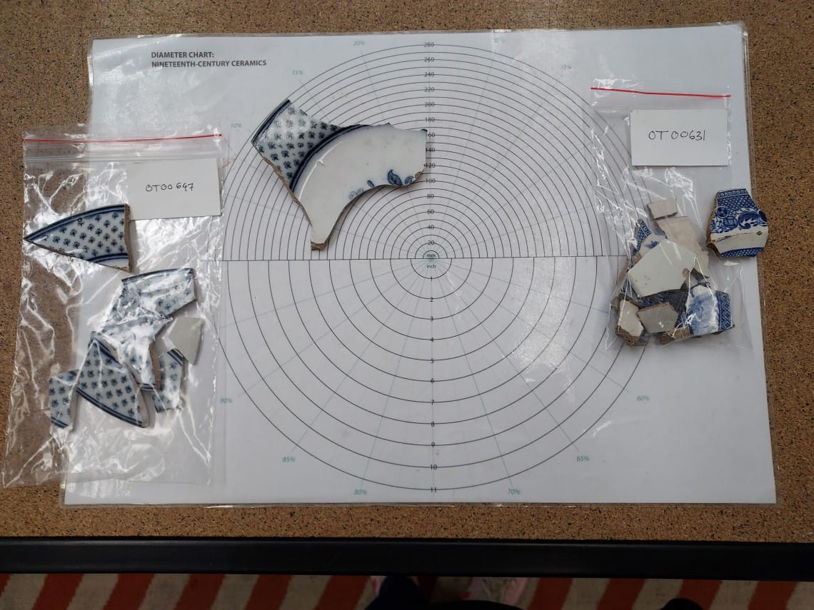A table with a piece of paper marking the outline of a plate. Fragments of pottery are being laid onto the outline to reconstruct a plate.